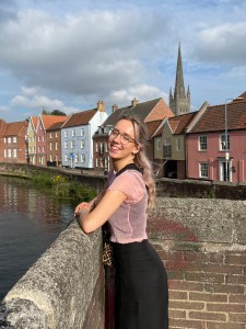 Aimée Rumsby, a blonde woman, standing at Rye Bridge in Norwich. Behind Aimée is Norwich Cathedral and a row of colourful homes.